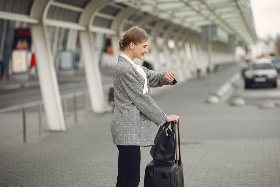 Side view of happy woman in checkered jacket standing with luggage and looking on watch before flight departure in airport