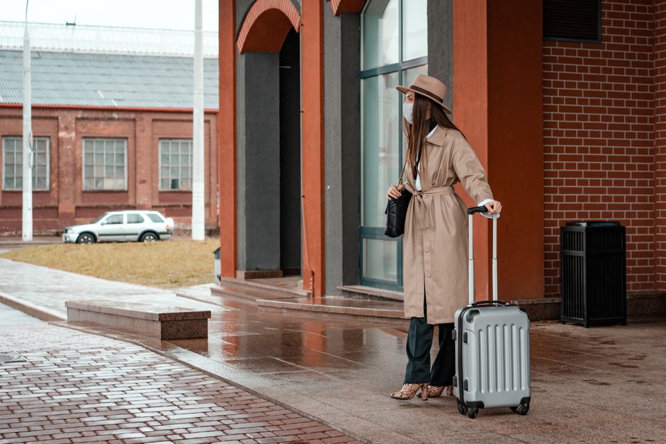 Fashionable woman in beige coat and hat with suitcase standing outdoors.