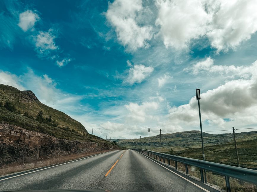 Open road through rugged mountains under a bright blue sky with clouds.
