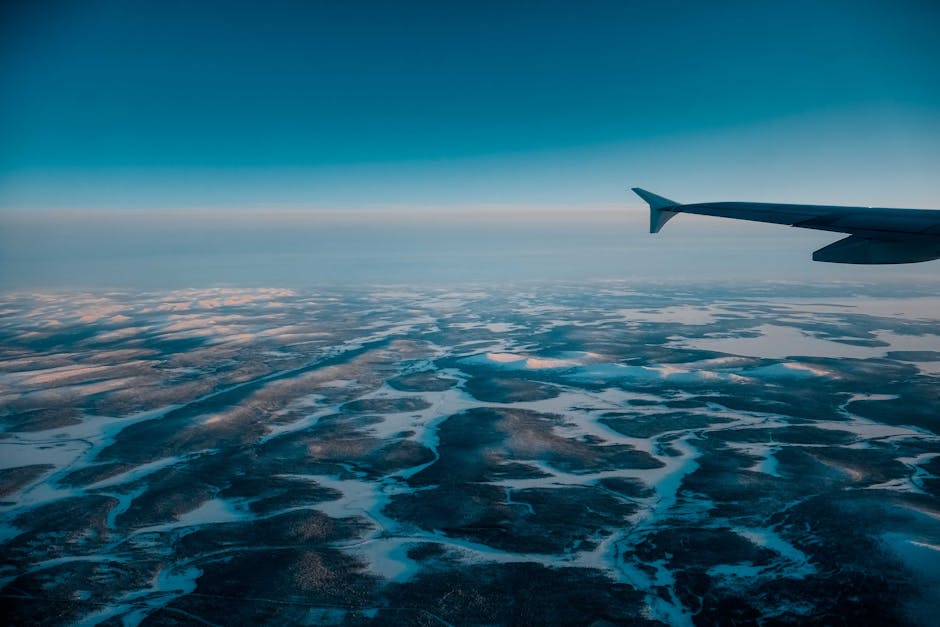 Wing of airplane flying in blue cloudless sky with skyline above vast terrain covered with snow in nature on winter day