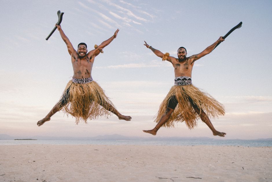 Joyful Fijian men in traditional skirts leaping on a beach with the ocean in the background.