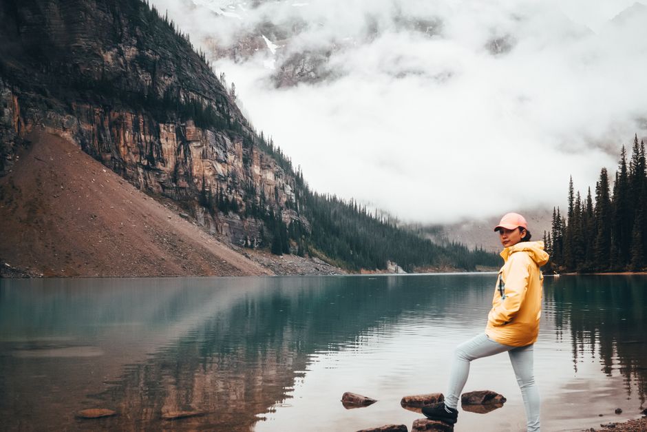 A woman in a yellow jacket stands by a serene, misty mountain lake, capturing a sense of adventure.