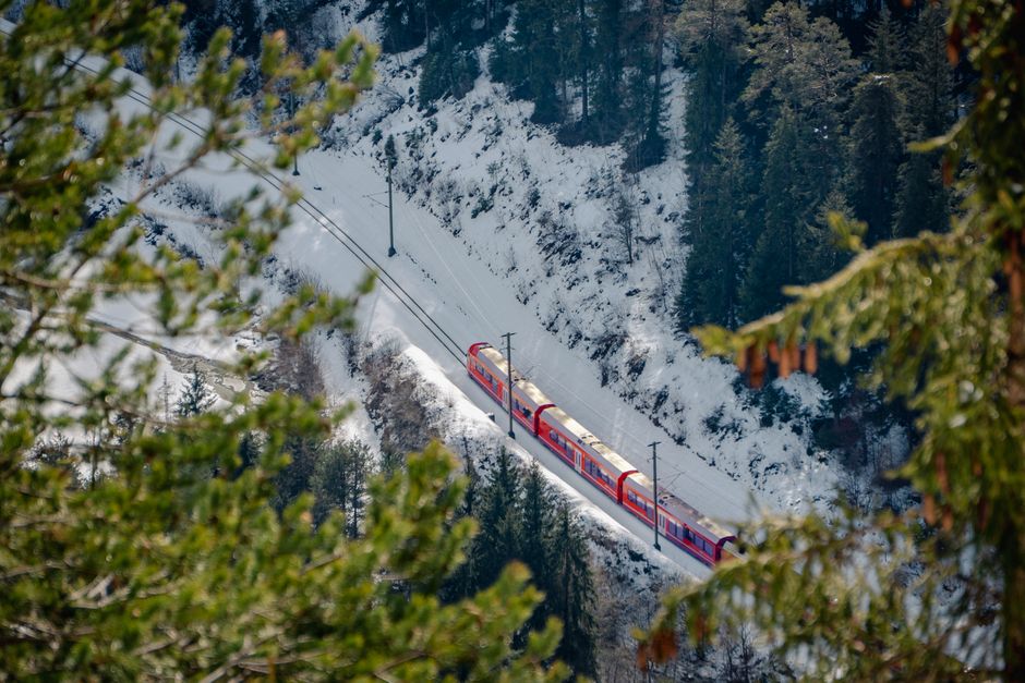 A red train traverses snowy mountains surrounded by pine trees, capturing a serene winter landscape.