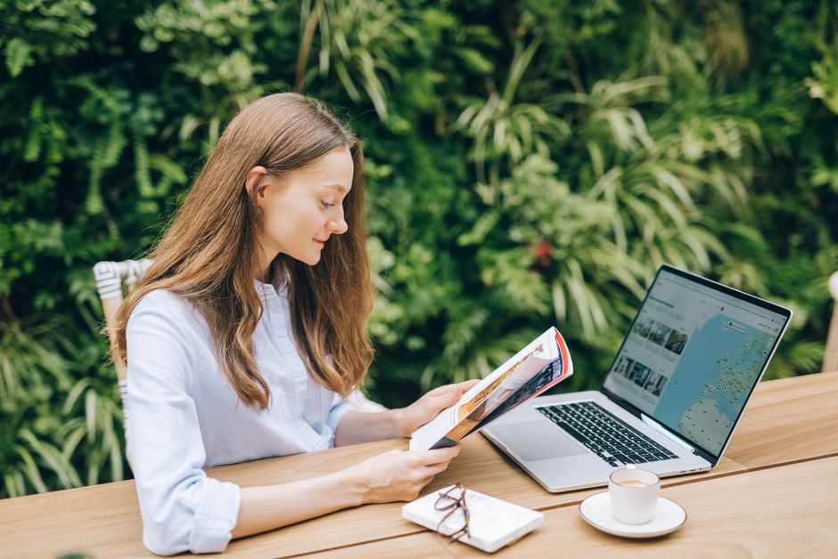 Caucasian woman reading a magazine at a wooden table with a laptop, outdoors.