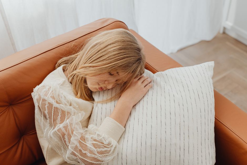 A young girl with blonde hair sleeps peacefully on a sofa, resting her head on a striped pillow indoors.