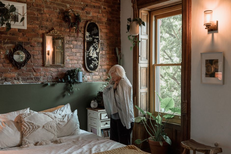 Elderly woman standing in a cozy bedroom with brick walls and soft lighting.