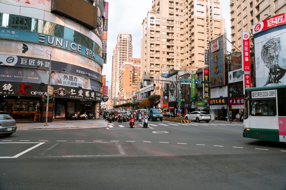 Busy urban intersection in Taipei, with scooters and buses under towering buildings.